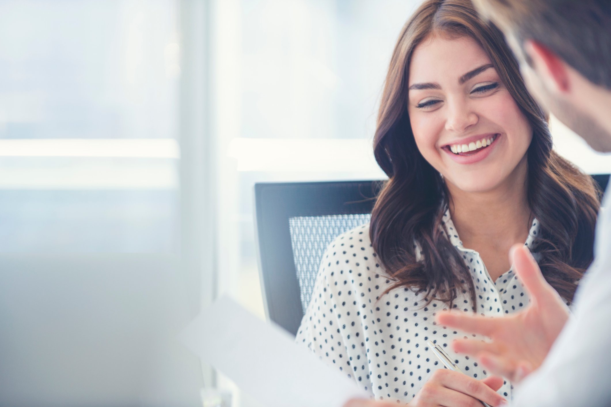 Two colleagues working together at a table in a well lit meeting room. While holding paper, they are in conversation laughing.