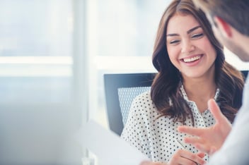 Two colleagues working together at a table in a well lit meeting room. While holding paper, they are in conversation laughing.