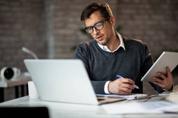 man holding a pen and tablet looking at his laptop