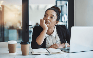 Woman sitting in front of computer staring off into space with open notebook and two cups of coffee on the desk.
