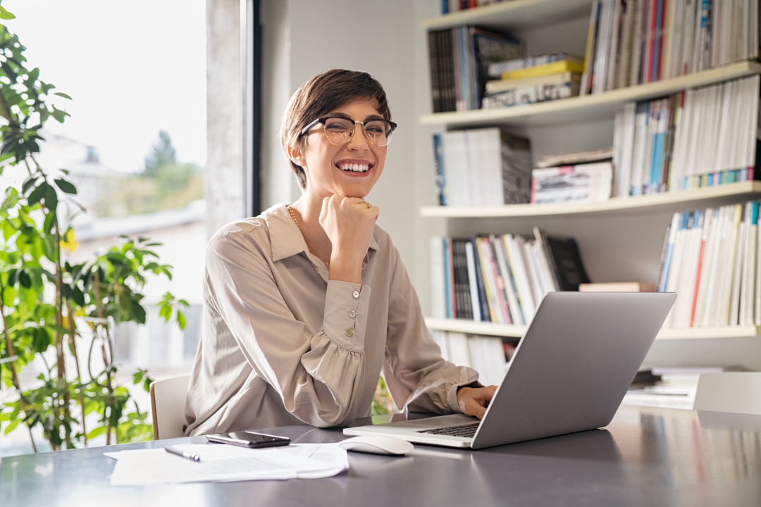 Successful bookkeeper sitting in office and looking at camera, with hand on chin working on computer while smiling
