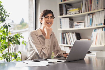 Successful bookkeeper sitting in office and looking at camera, with hand on chin working on computer while smiling