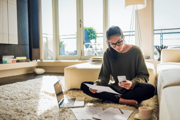 Young person sitting on carpet next to a sofa with paper and mobile phone in hand. There is a laptop and tea cup on the carpet.